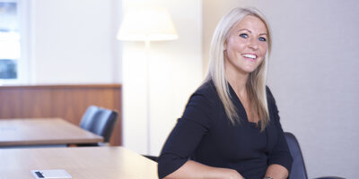 Lady with blonde hair smiling at camera , seated with her elbow on desk.